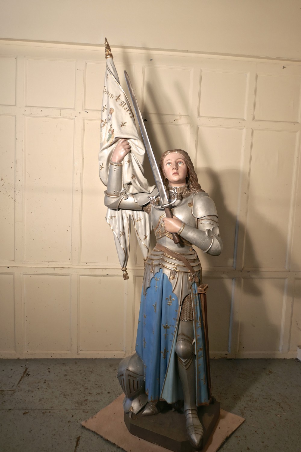 A French polychrome plaster statue depicting Saint Joan of Arc in silver armour and a blue tunic decorated with fleur-de-lis, holding a sword and flag. View IV