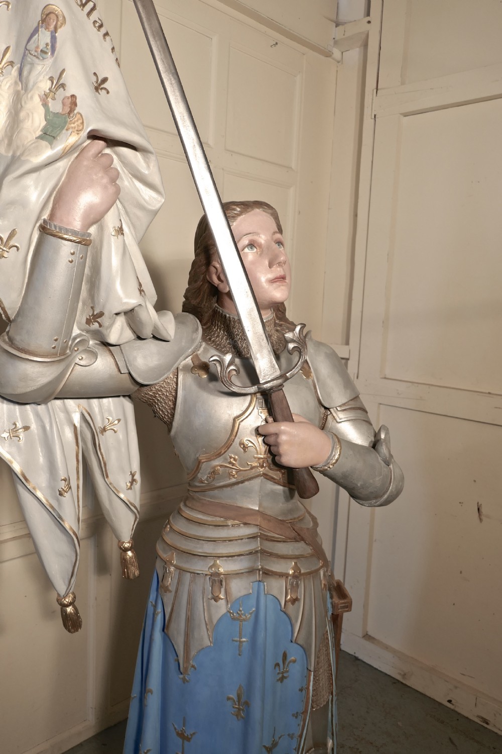 A French polychrome plaster statue depicting Saint Joan of Arc in silver armour and a blue tunic decorated with fleur-de-lis, holding a sword and flag. View VIII