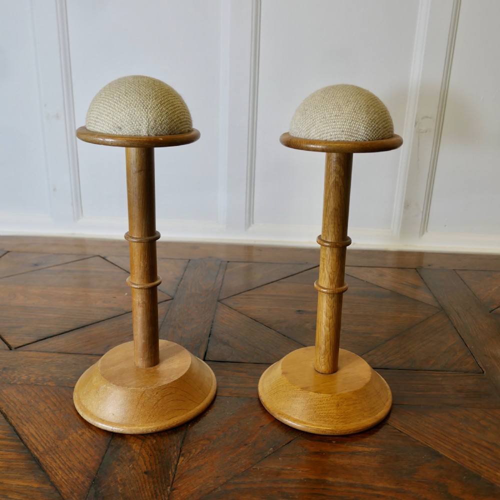 A pair of turned golden oak hat stands featuring circular weighted bases and woven fabric upholstered dome tops.