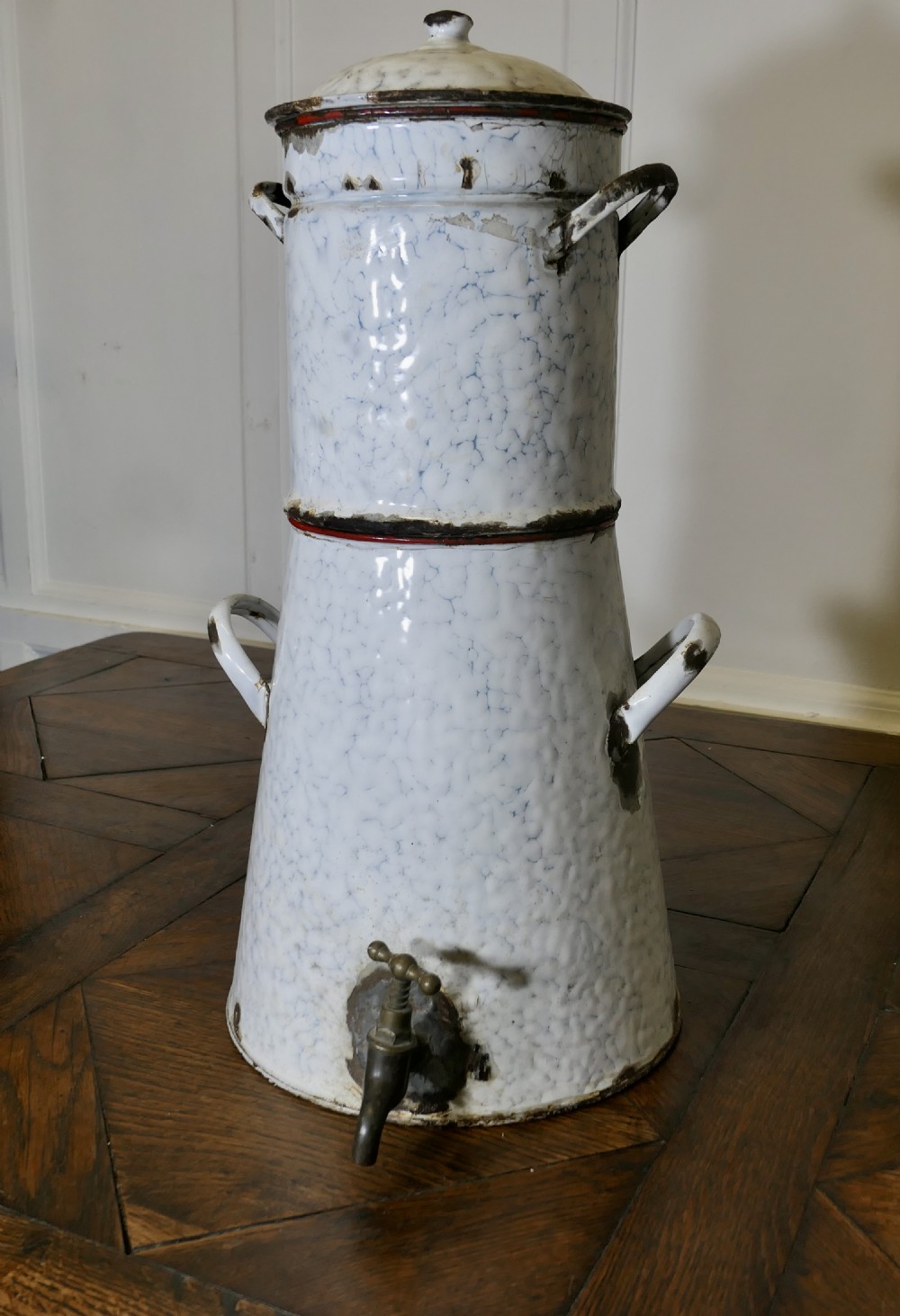 A tall antique white and blue marbled enamel coffee urn featuring a brass tap near the base, loop side handles, and red trim along the rim.