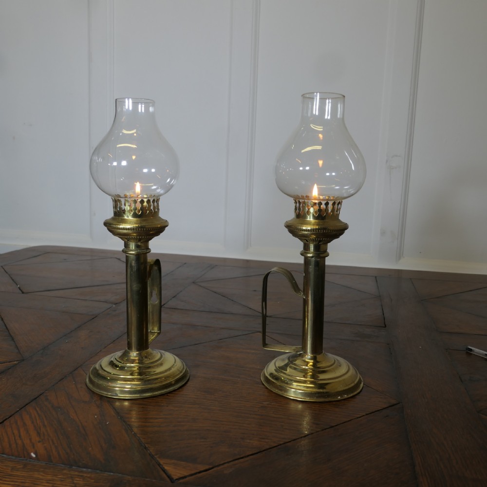 A pair of polished brass candlesticks featuring side handles and removable clear glass hurricane chimneys enclosing lit candles.