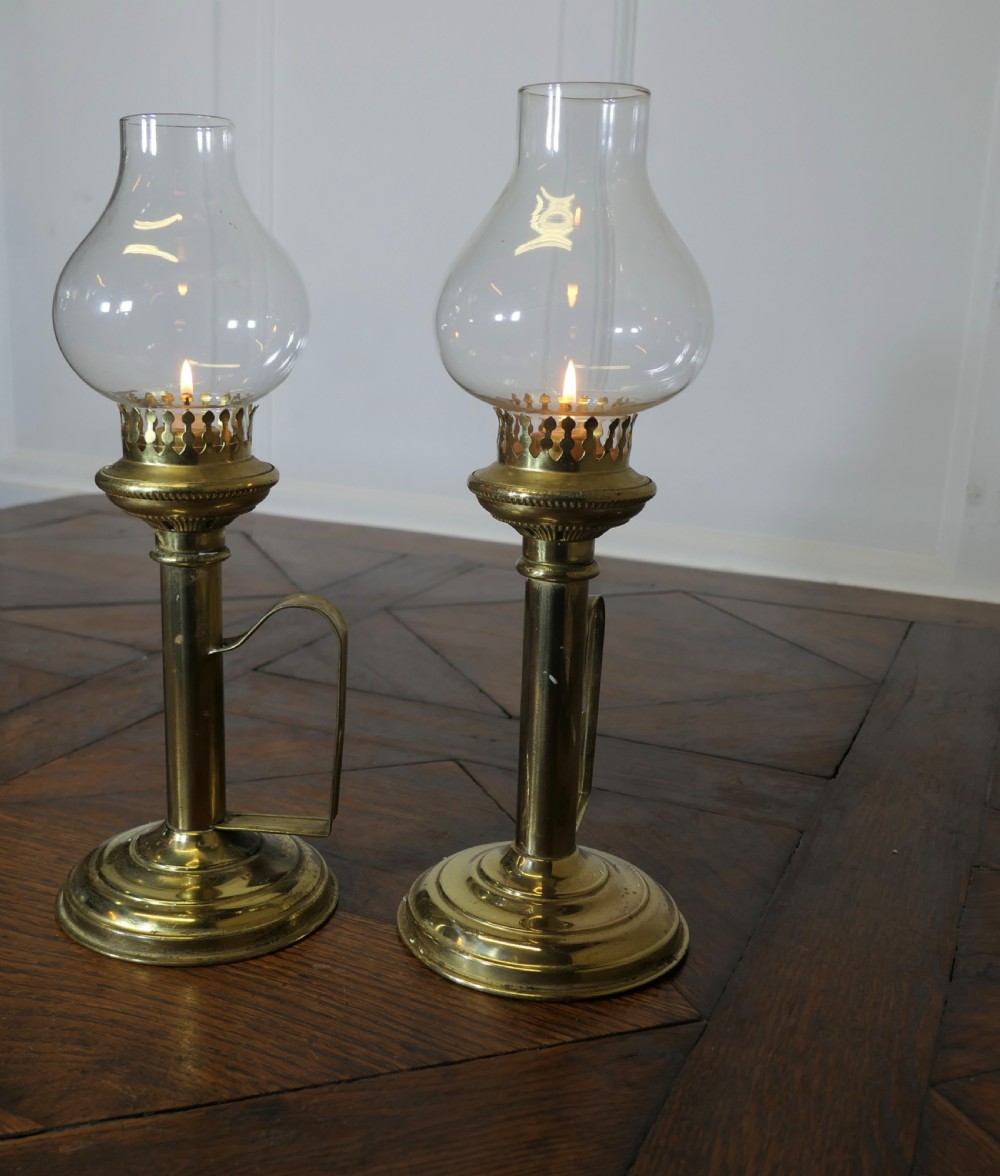 A pair of polished brass candlesticks featuring side handles and removable clear glass hurricane chimneys enclosing lit candles. View II