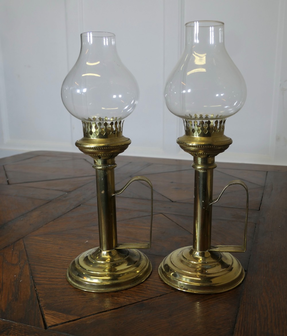 A pair of polished brass candlesticks featuring side handles and removable clear glass hurricane chimneys enclosing lit candles. View V