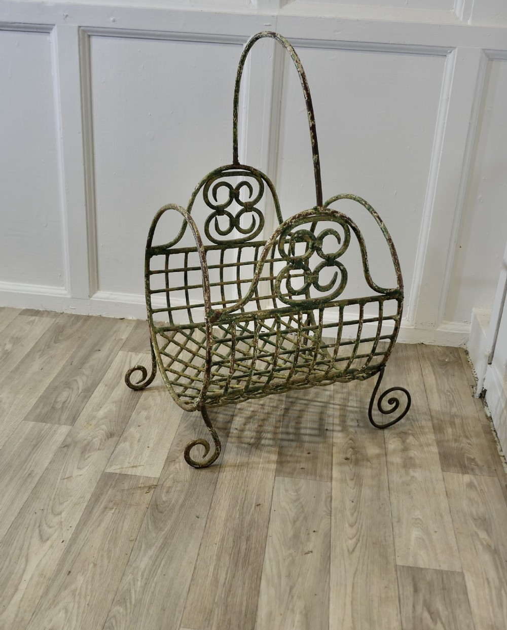 A 19th-century French distressed green painted wrought iron wirework log carrier featuring scroll feet, an elevated loop handle, and open mesh grid sides.