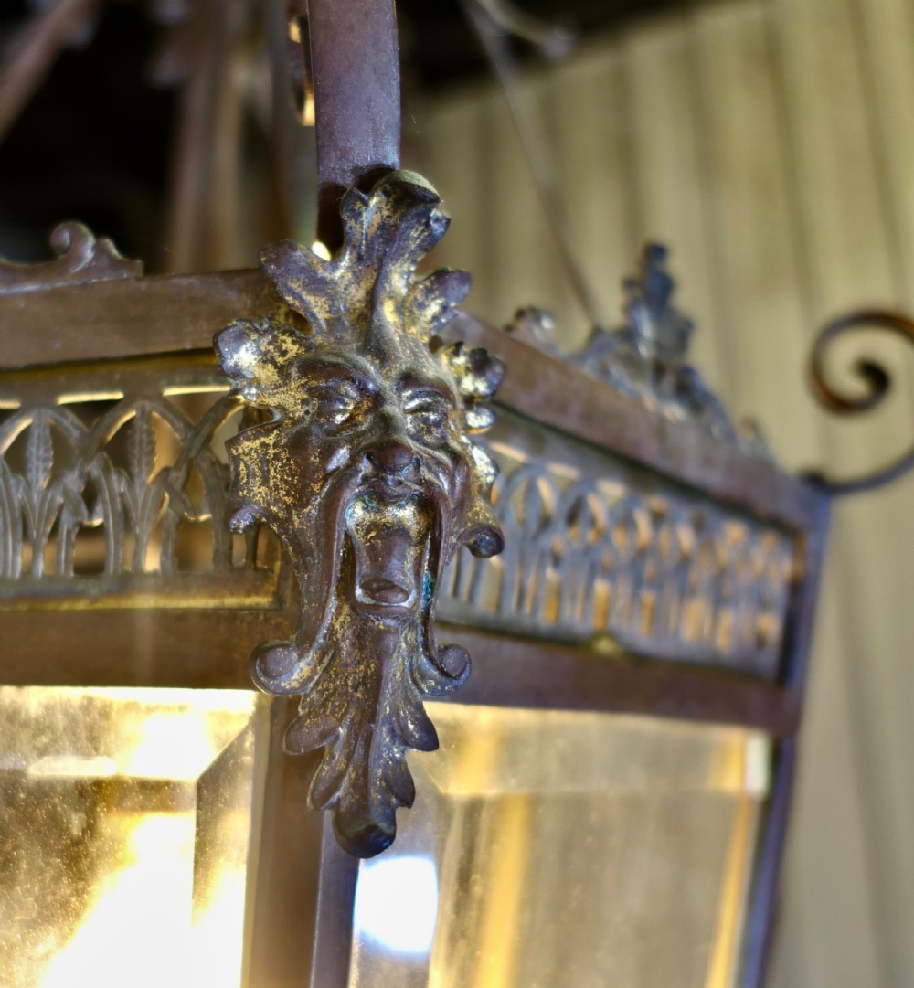 A dark patinated copper hexagonal ceiling lantern with glazed glass panels, scrolled metal supports, and cast Green Man mask decorations. View V