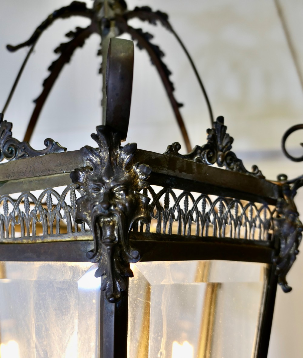 A dark patinated copper hexagonal ceiling lantern with glazed glass panels, scrolled metal supports, and cast Green Man mask decorations. View VI