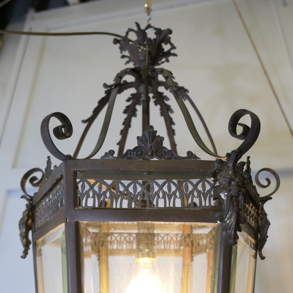 A dark patinated copper hexagonal ceiling lantern with glazed glass panels, scrolled metal supports, and cast Green Man mask decorations. View VII