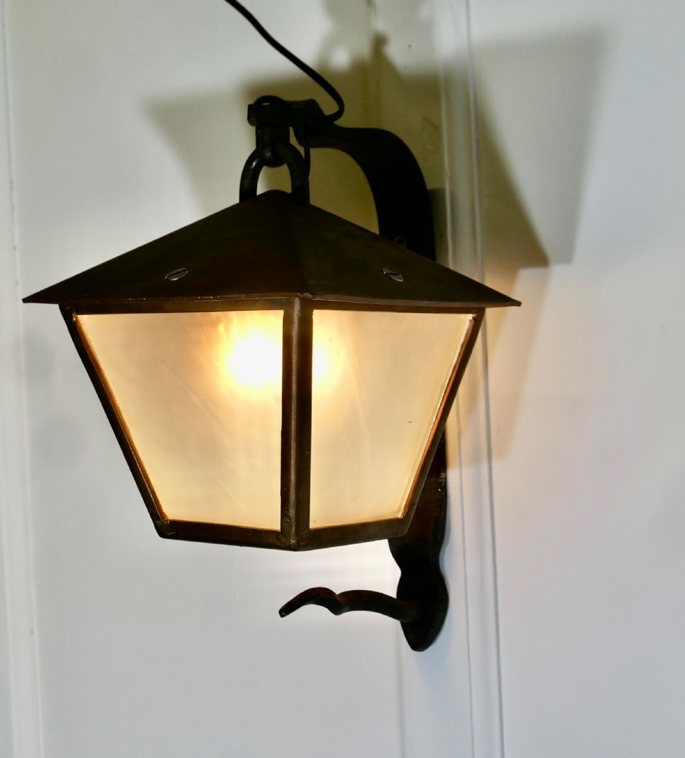 A wrought iron and copper wall lantern featuring a gooseneck bracket and frosted glass panels illuminated from within. View VI