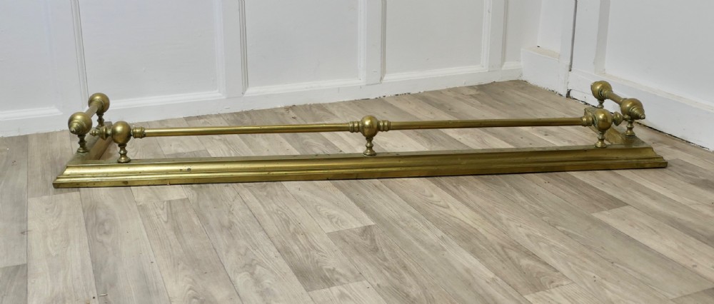 A large Victorian brass fireplace fender featuring a low moulded base topped with turned brass finials supporting a horizontal rail.