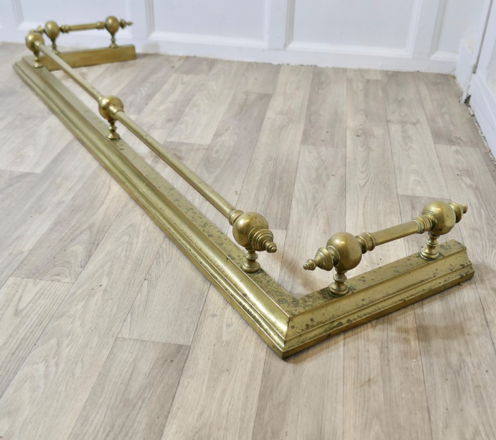 A large Victorian brass fireplace fender featuring a low moulded base topped with turned brass finials supporting a horizontal rail. View V