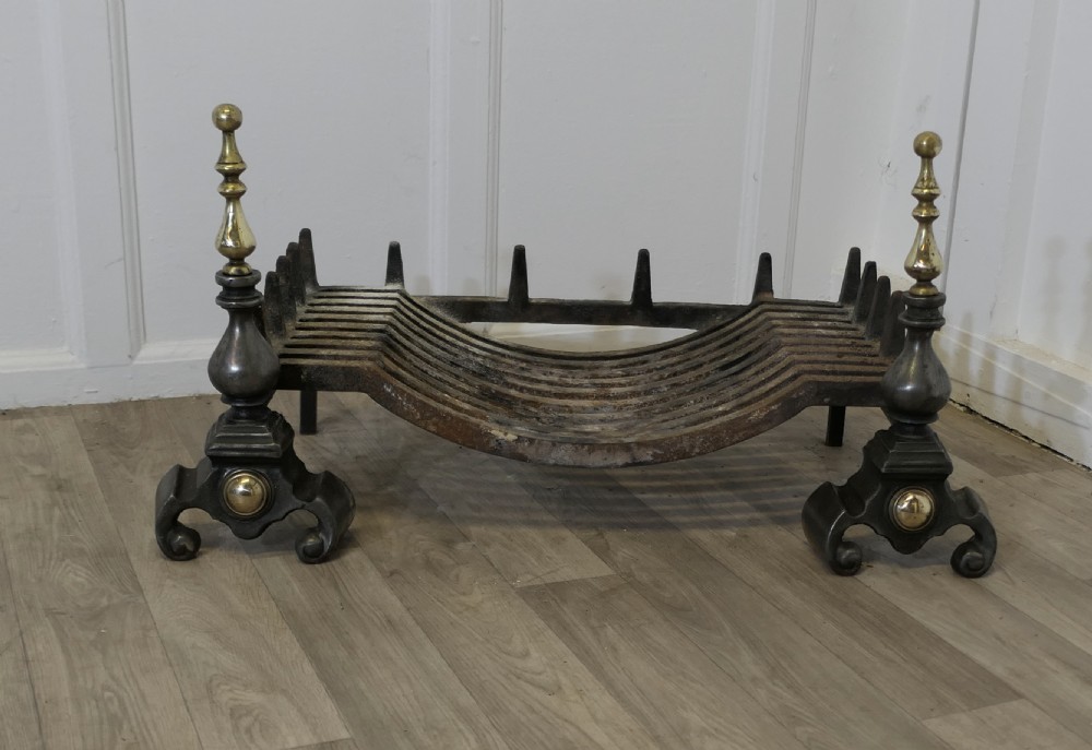A heavy curved steel log grate supported by two front andirons featuring scroll feet, central brass cabochons, and turned brass urn finials.