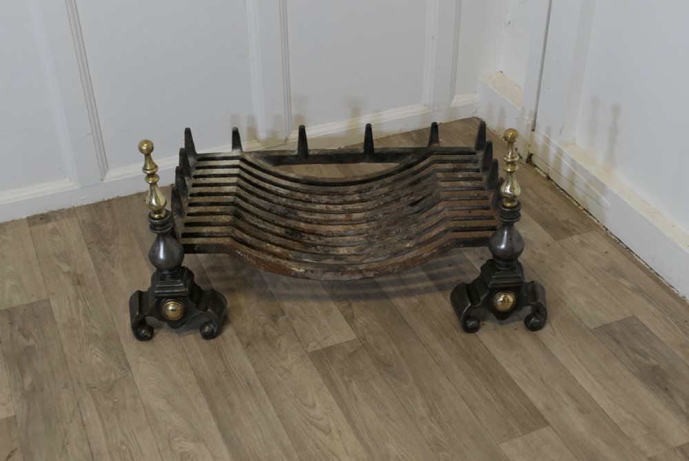 A heavy curved steel log grate supported by two front andirons featuring scroll feet, central brass cabochons, and turned brass urn finials. View III