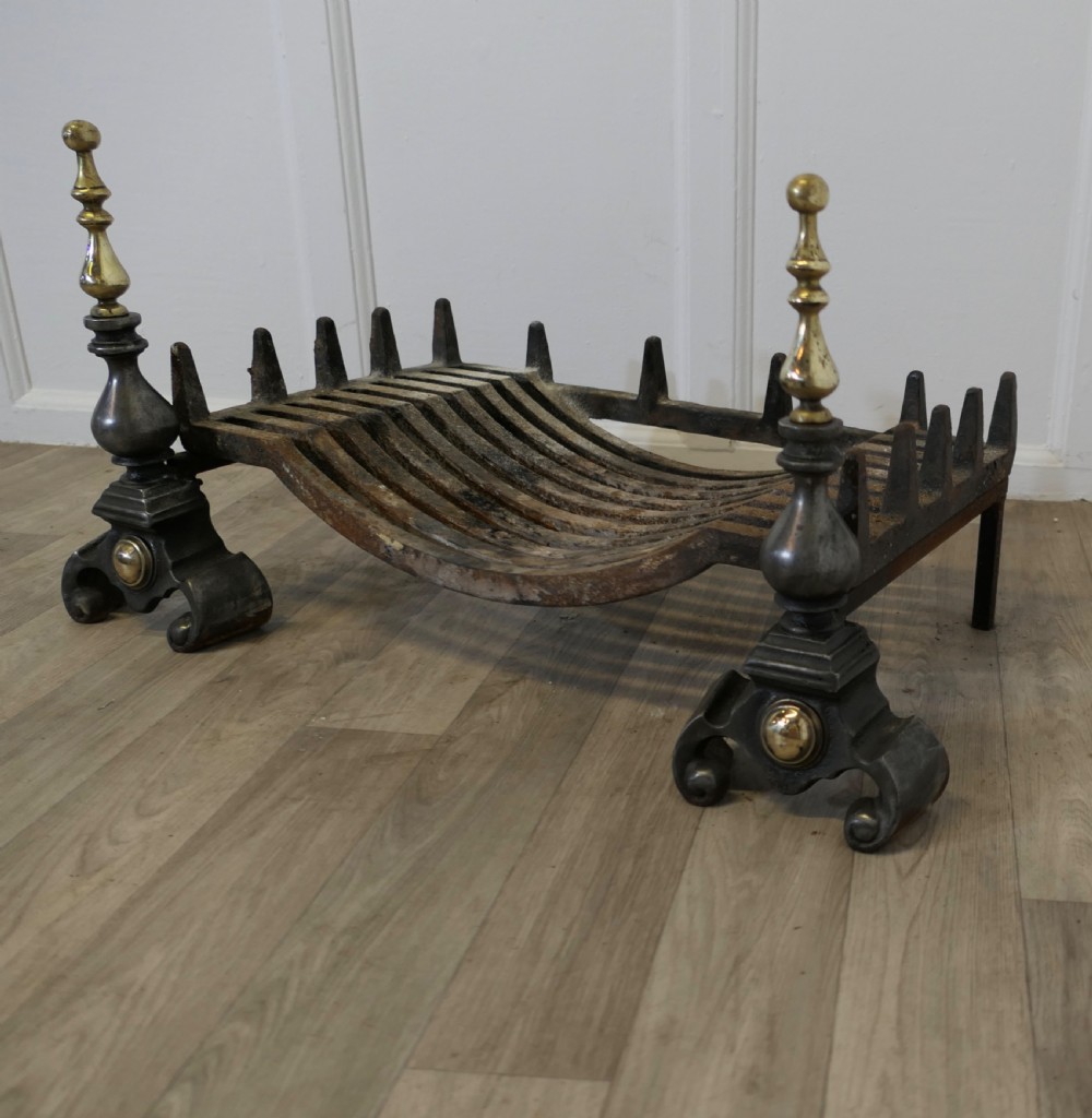A heavy curved steel log grate supported by two front andirons featuring scroll feet, central brass cabochons, and turned brass urn finials. View IV