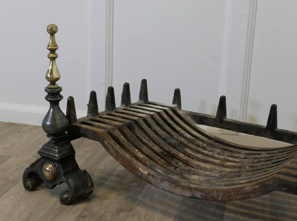 A heavy curved steel log grate supported by two front andirons featuring scroll feet, central brass cabochons, and turned brass urn finials. View V