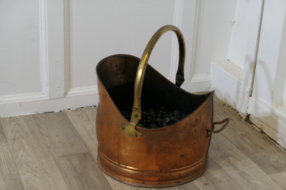 An Arts and Crafts beaten copper helmet coal scuttle featuring a brass overhead swing handle and a small rear tipping handle.