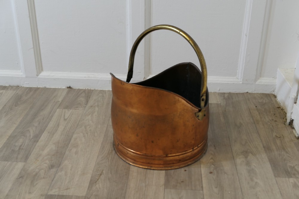 An Arts and Crafts beaten copper helmet coal scuttle featuring a brass overhead swing handle and a small rear tipping handle. View V