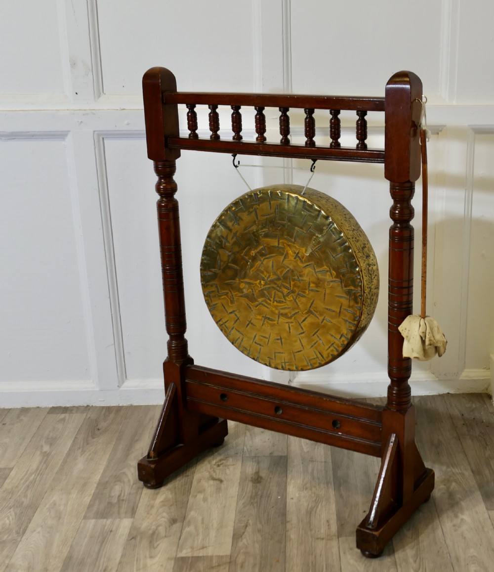 An Arts and Crafts walnut floor-standing dinner gong with turned side columns, a spindle gallery, and a central circular hand-hammered brass disc with attached striker. View V