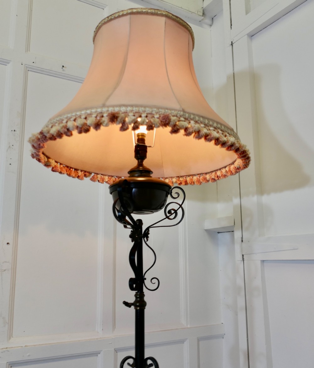 An antique wrought iron telescopic standard lamp featuring decorative scrollwork legs, a brass adjustment collar, an oil font mount, and a fringed fabric lampshade. View VI