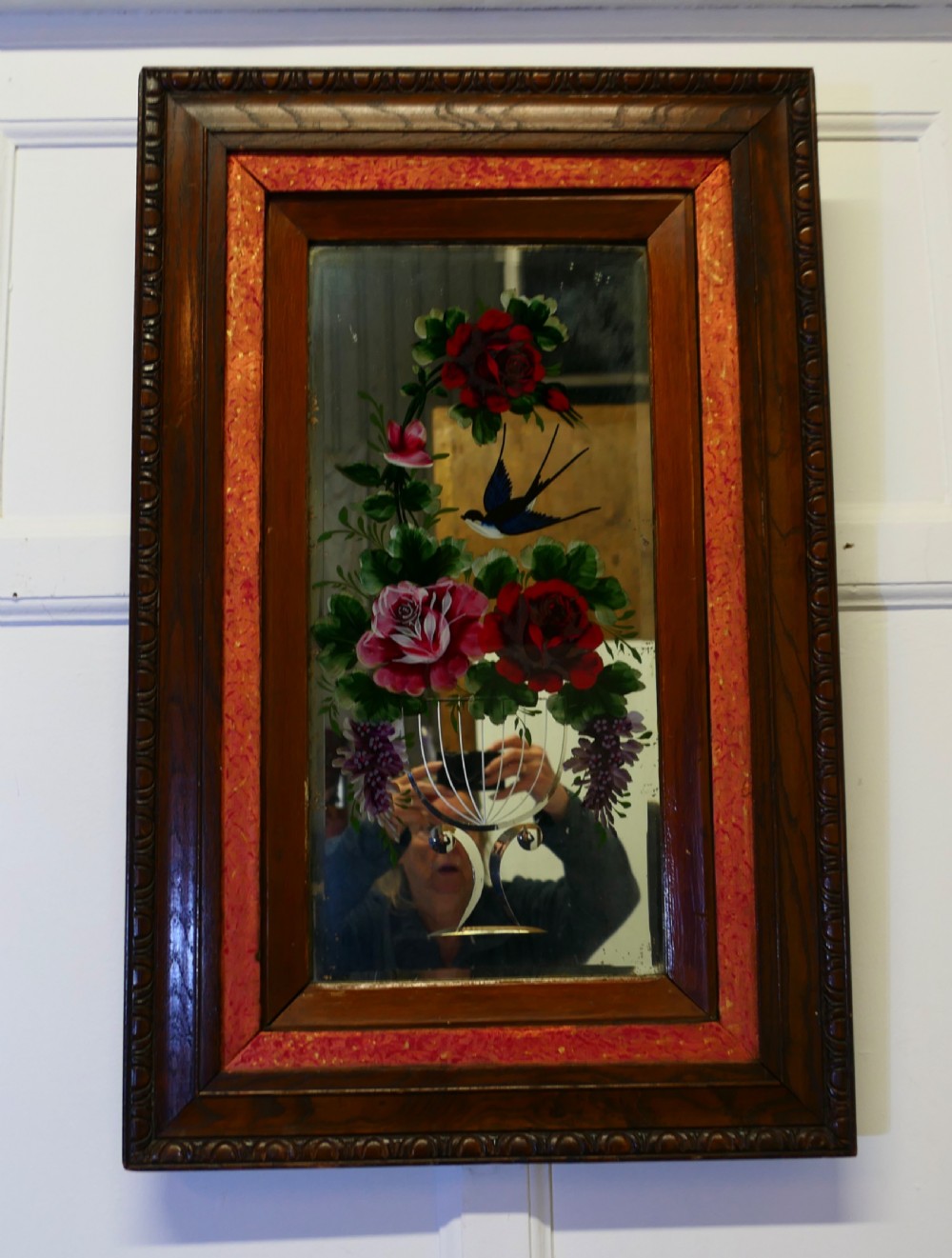 A rectangular Victorian wall mirror featuring a carved wooden frame, a red border, and a central reverse-painted glass panel displaying red roses and a bird in a vase.