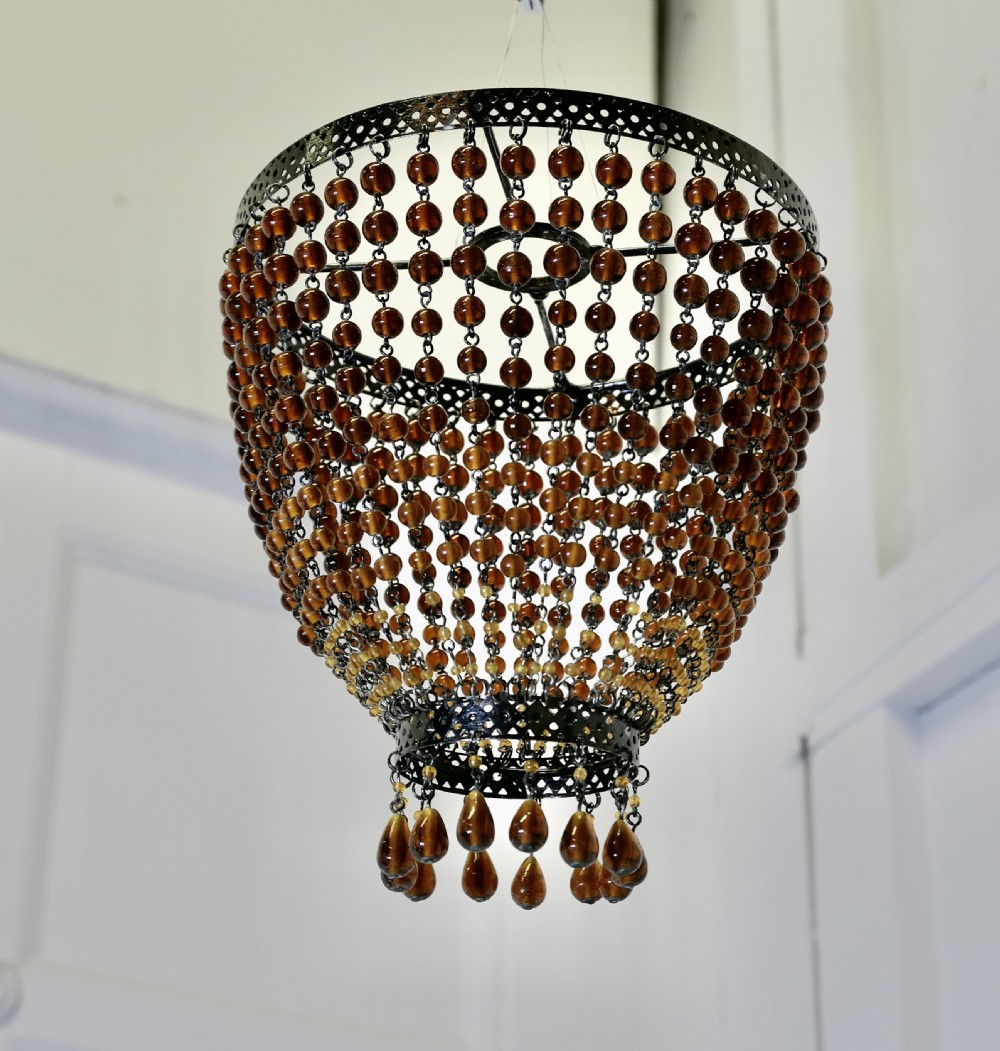 A basket-shaped lampshade crafted from amber glass beads strung together on a dark metal frame with dangling teardrop glass drops at the base. View II