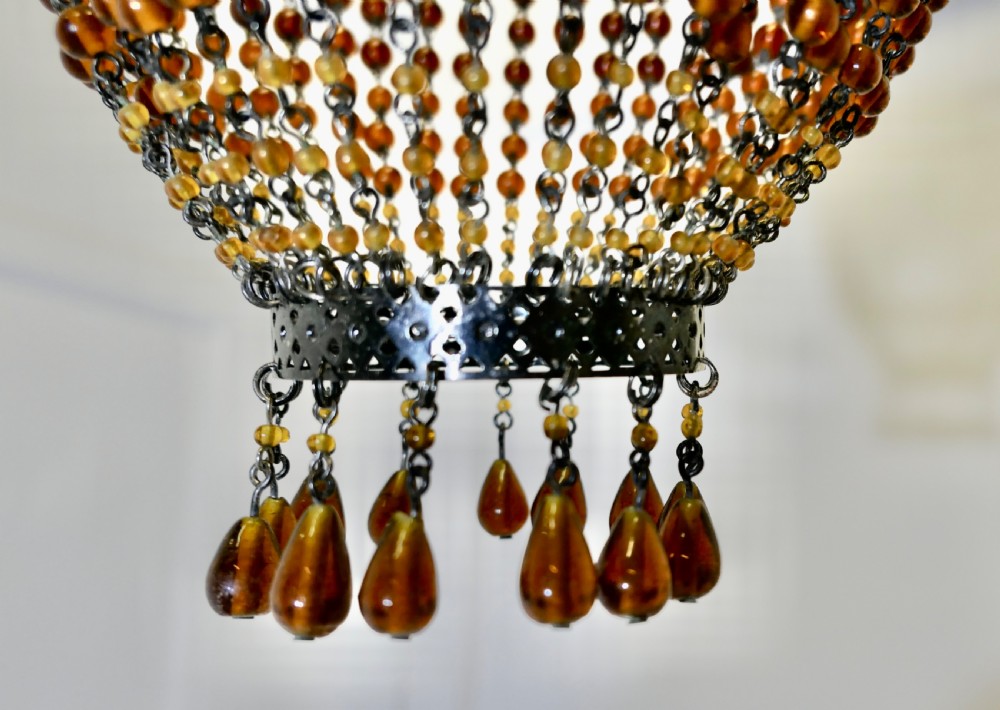 A basket-shaped lampshade crafted from amber glass beads strung together on a dark metal frame with dangling teardrop glass drops at the base. View IV