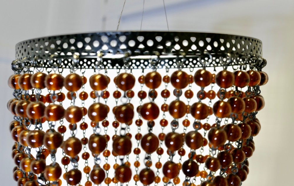 A basket-shaped lampshade crafted from amber glass beads strung together on a dark metal frame with dangling teardrop glass drops at the base. View V