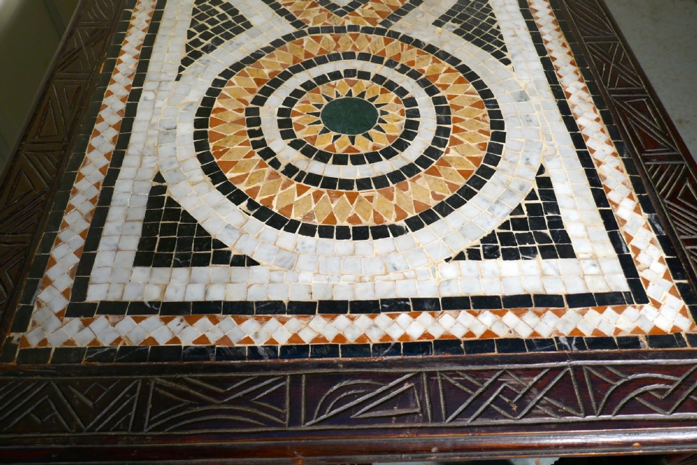 A rectangular dark wood dining table featuring a detailed geometric stone mosaic top, hand-carved geometric patterns on the apron, and carved legs connected by stretchers. View VI