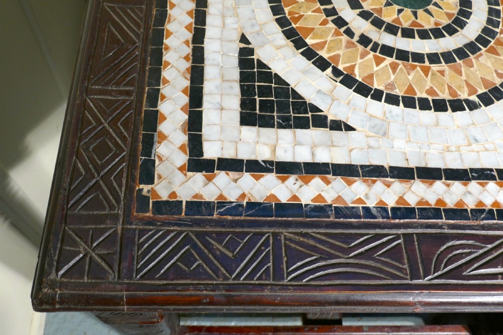 A rectangular dark wood dining table featuring a detailed geometric stone mosaic top, hand-carved geometric patterns on the apron, and carved legs connected by stretchers. View VII