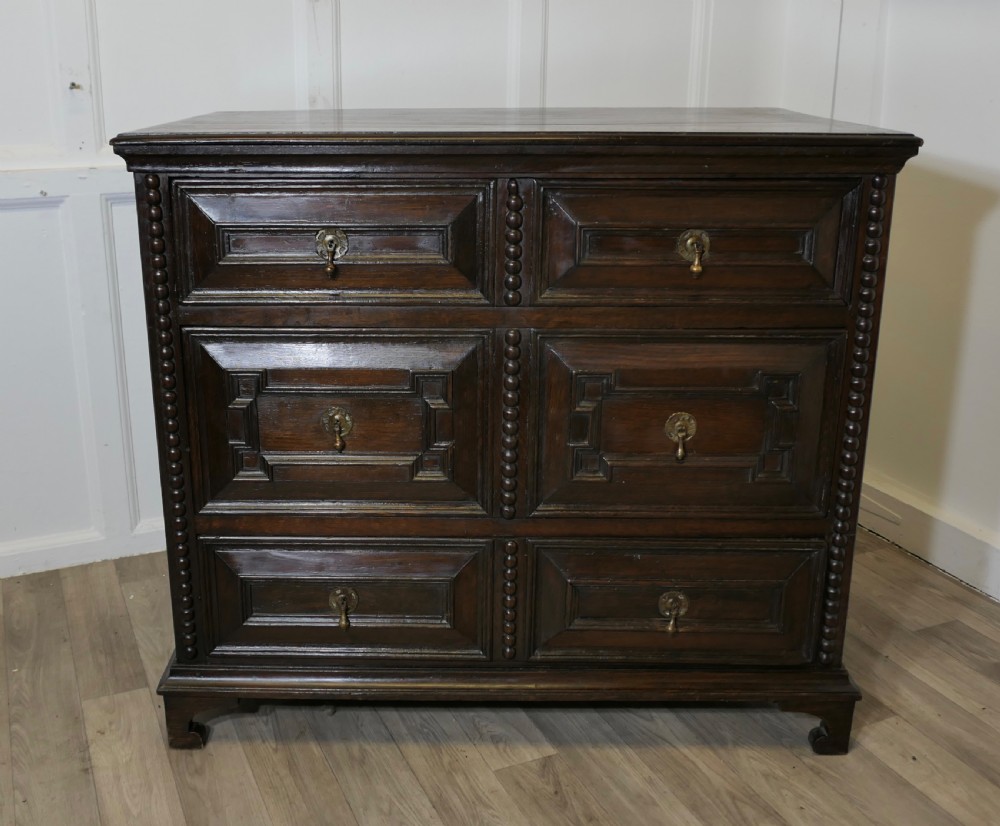 A dark oak chest of drawers featuring geometric moulded drawer fronts, vertical beaded moulding, and brass tear-drop handles.