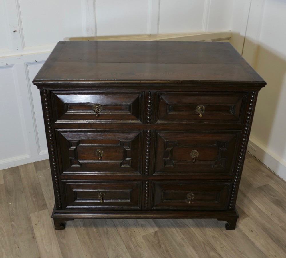 A dark oak chest of drawers featuring geometric moulded drawer fronts, vertical beaded moulding, and brass tear-drop handles. View II
