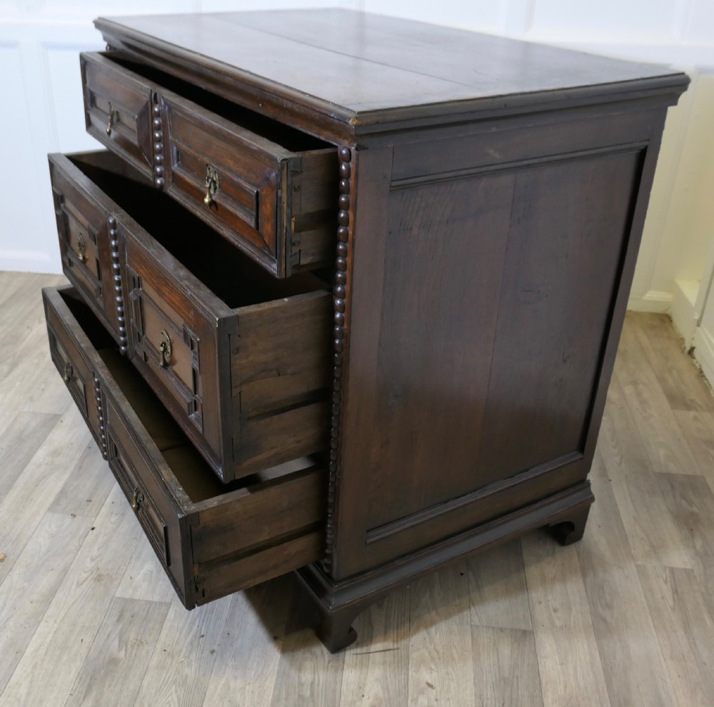 A dark oak chest of drawers featuring geometric moulded drawer fronts, vertical beaded moulding, and brass tear-drop handles. View V