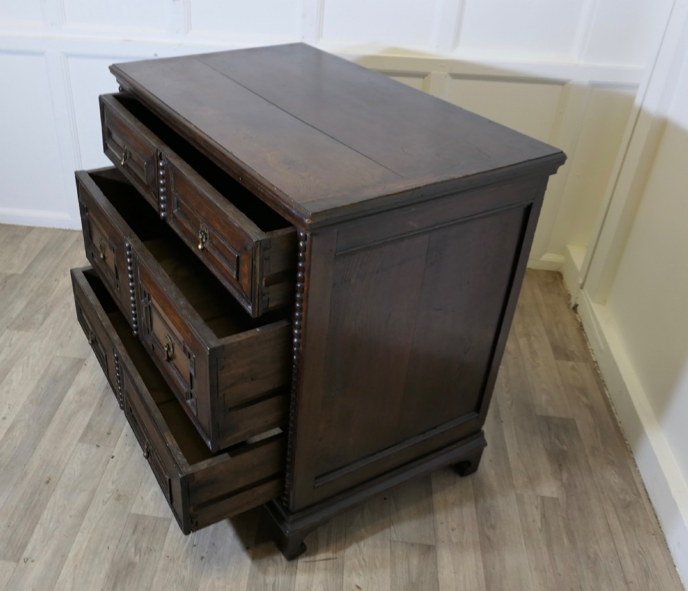 A dark oak chest of drawers featuring geometric moulded drawer fronts, vertical beaded moulding, and brass tear-drop handles. View VI