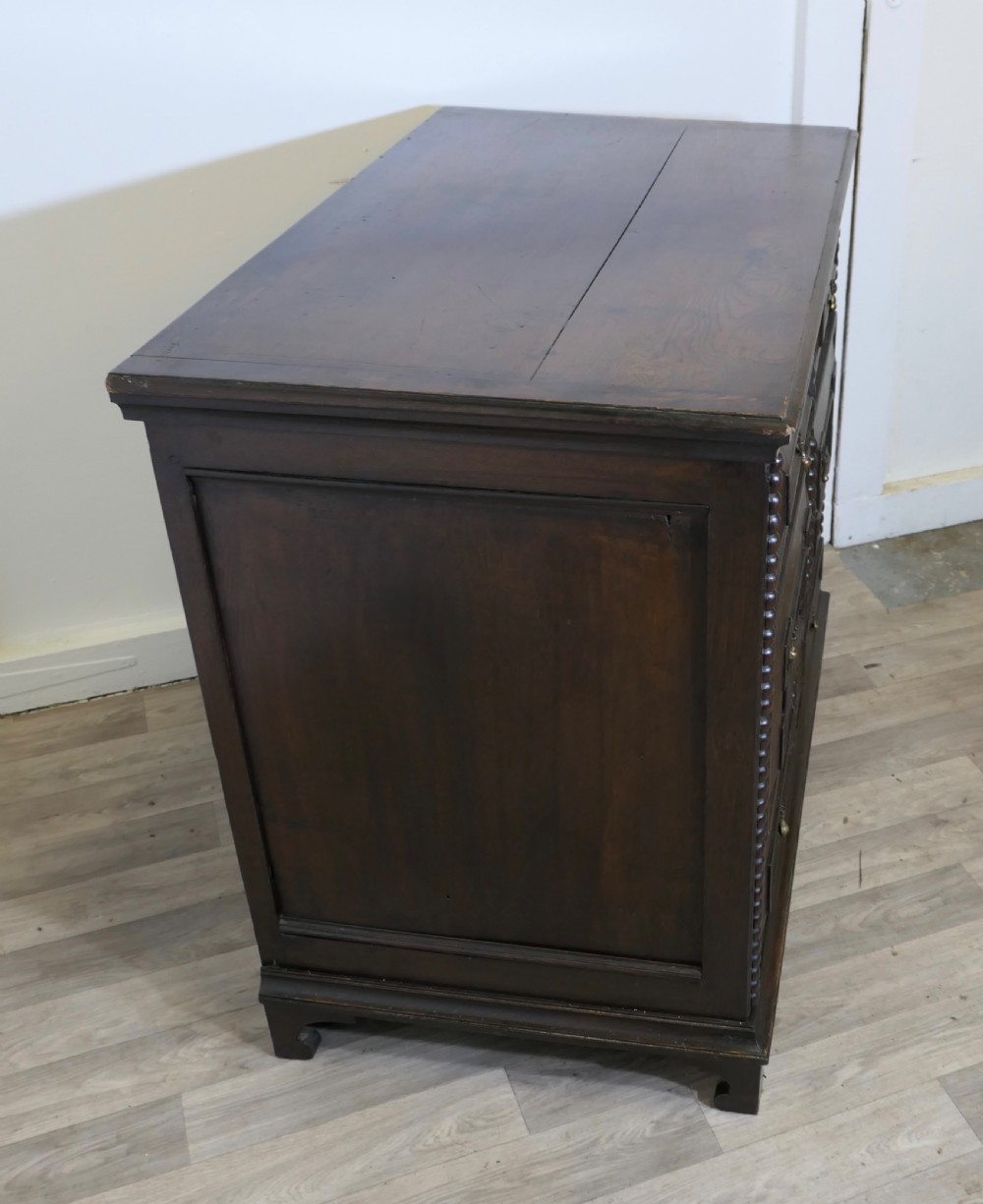 A dark oak chest of drawers featuring geometric moulded drawer fronts, vertical beaded moulding, and brass tear-drop handles. View VII