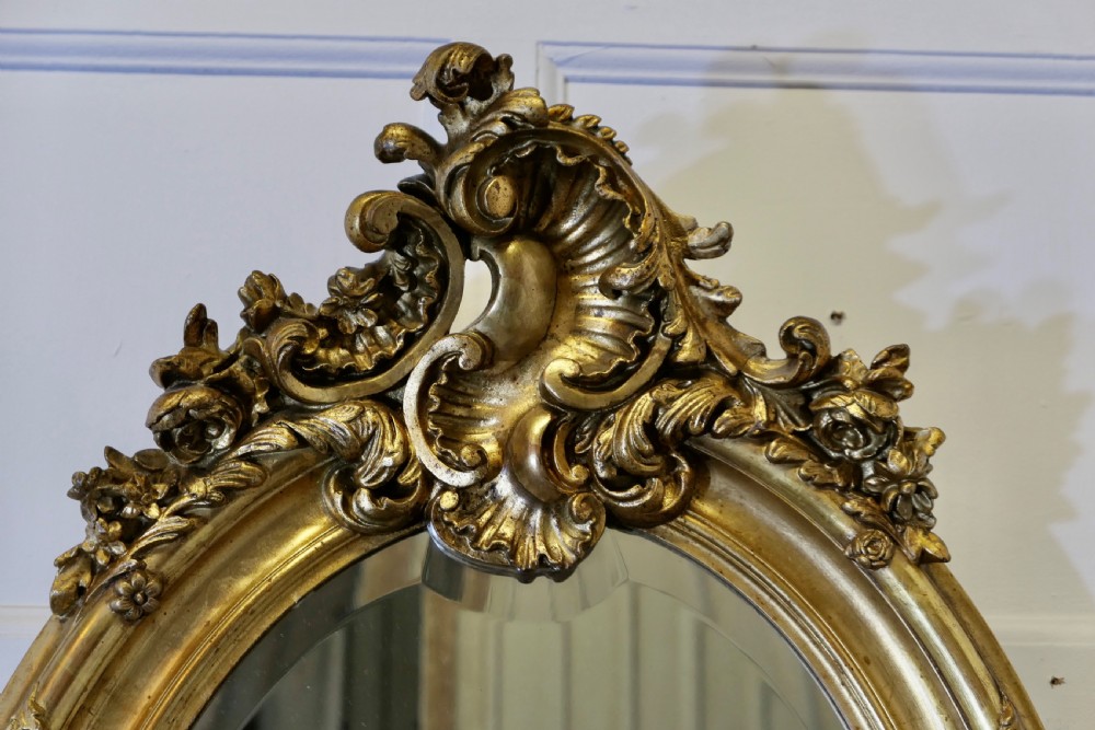 An oval wall mirror featuring an ornate gold gilded frame with elaborate top and bottom scrollwork cresting. View III