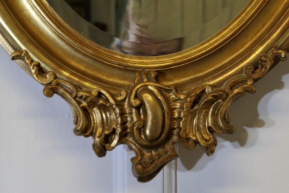 An oval wall mirror featuring an ornate gold gilded frame with elaborate top and bottom scrollwork cresting. View IV