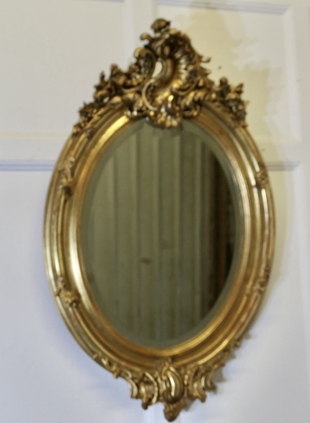 An oval wall mirror featuring an ornate gold gilded frame with elaborate top and bottom scrollwork cresting. View VI