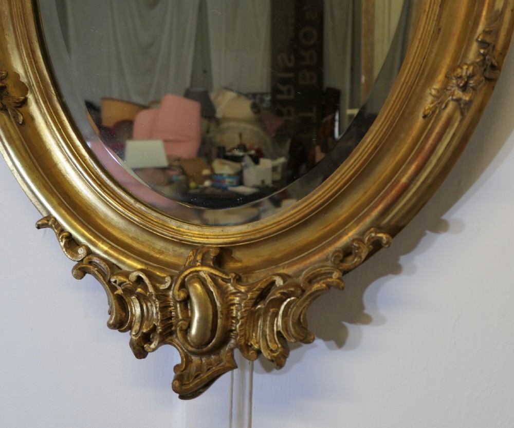An oval wall mirror featuring an ornate gold gilded frame with elaborate top and bottom scrollwork cresting. View VII