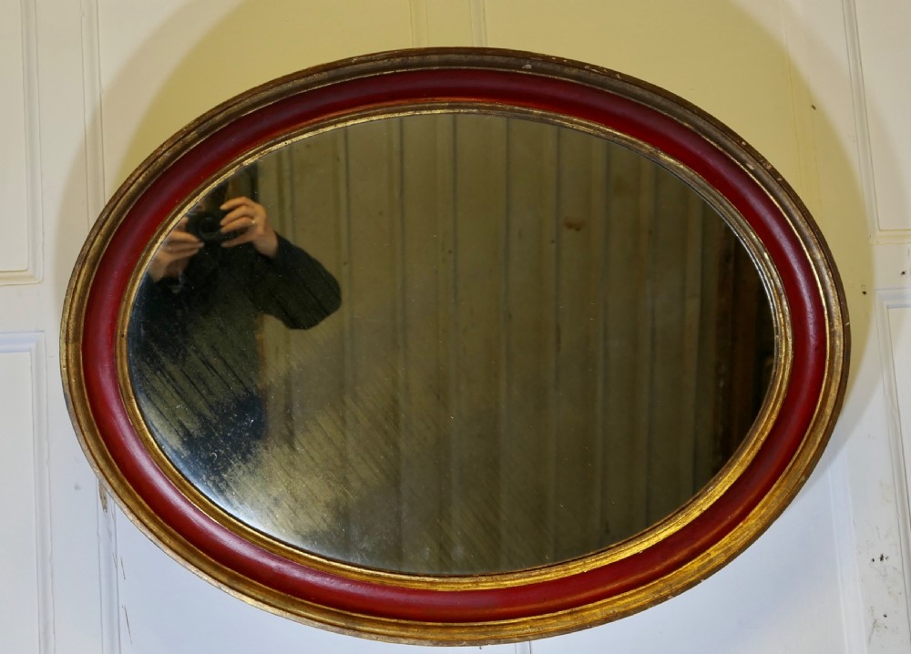 A large oval wall mirror featuring a deep red rounded moulding with gilded inner and outer borders.