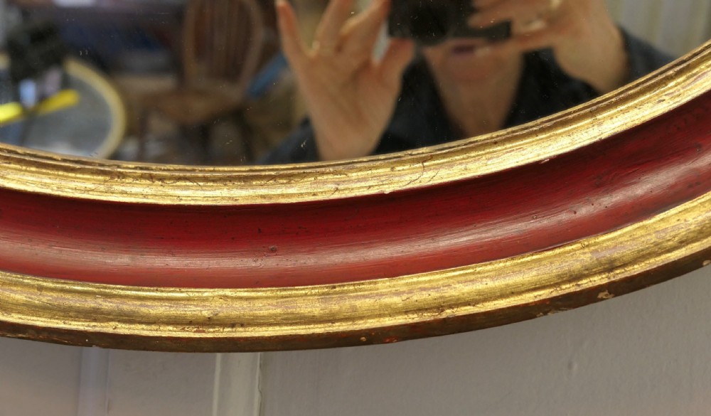 A large oval wall mirror featuring a deep red rounded moulding with gilded inner and outer borders. View VII