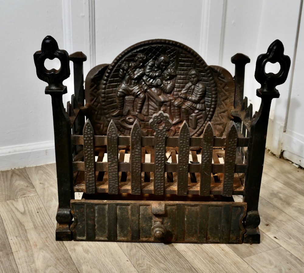 A heavy dark cast iron fire basket featuring a decorative relief backplate depicting figures, upright gothic finials, and a removable ash pan beneath.