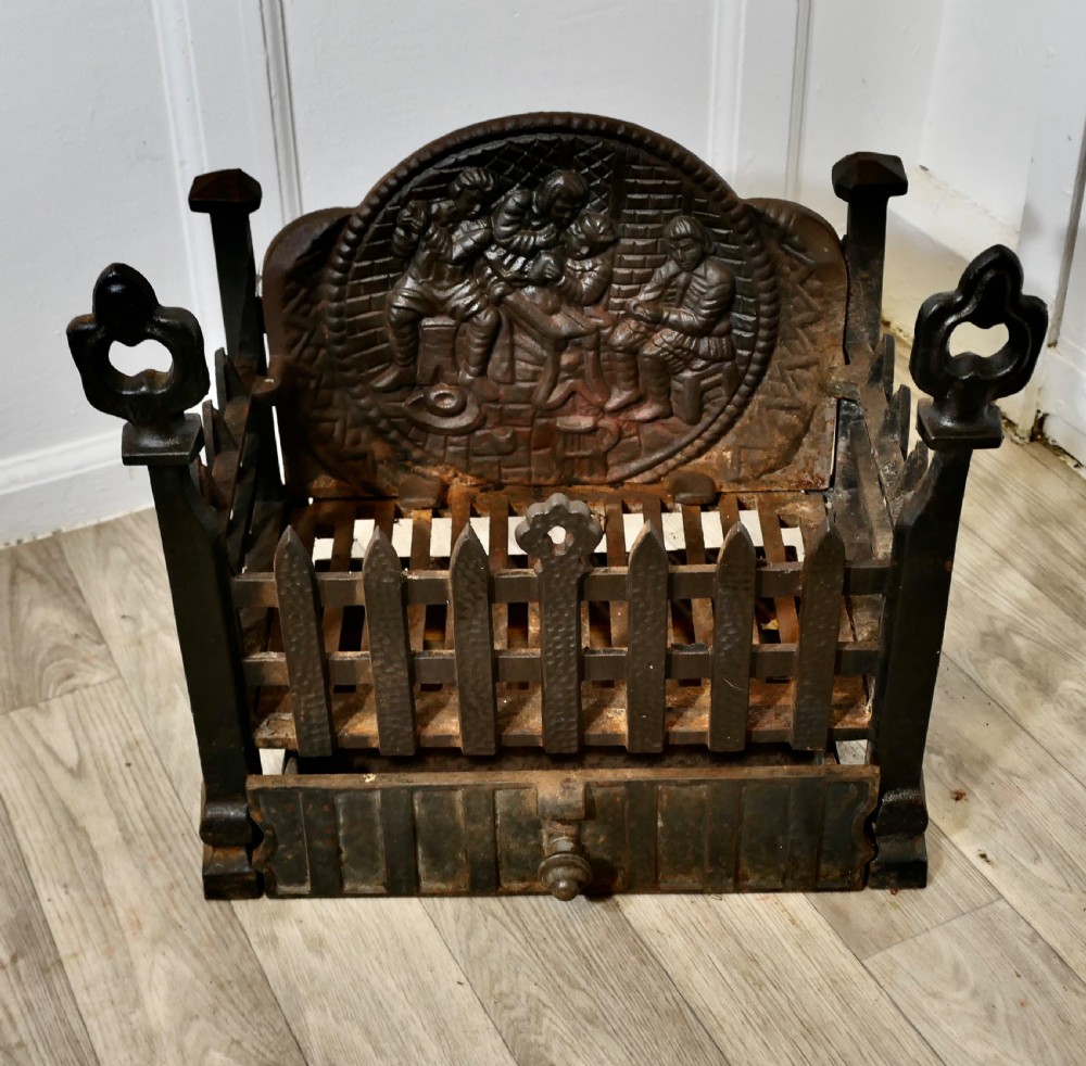 A heavy dark cast iron fire basket featuring a decorative relief backplate depicting figures, upright gothic finials, and a removable ash pan beneath. View II