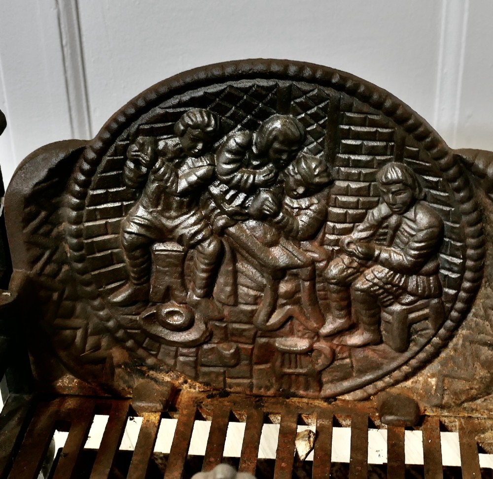 A heavy dark cast iron fire basket featuring a decorative relief backplate depicting figures, upright gothic finials, and a removable ash pan beneath. View III