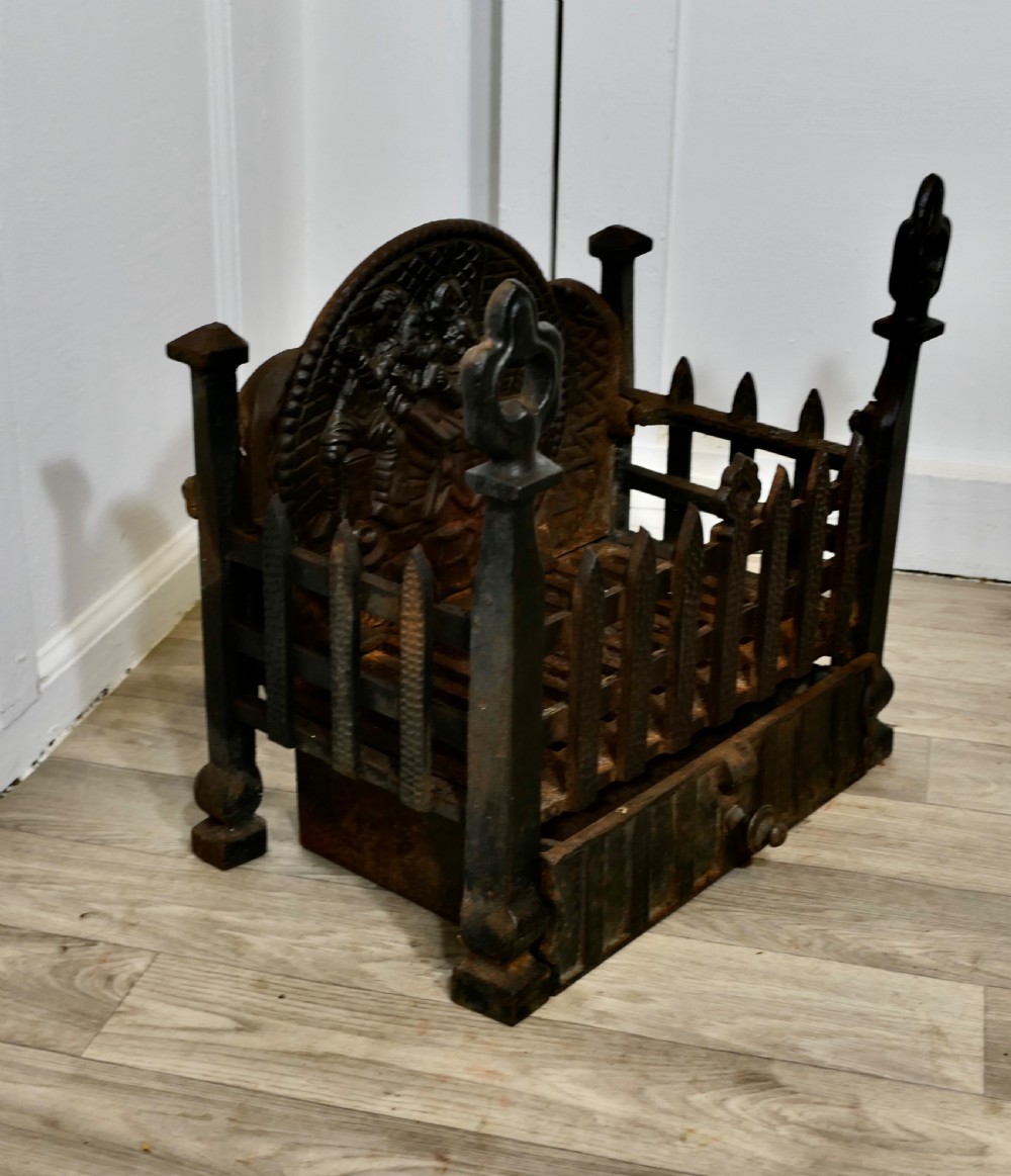 A heavy dark cast iron fire basket featuring a decorative relief backplate depicting figures, upright gothic finials, and a removable ash pan beneath. View IV