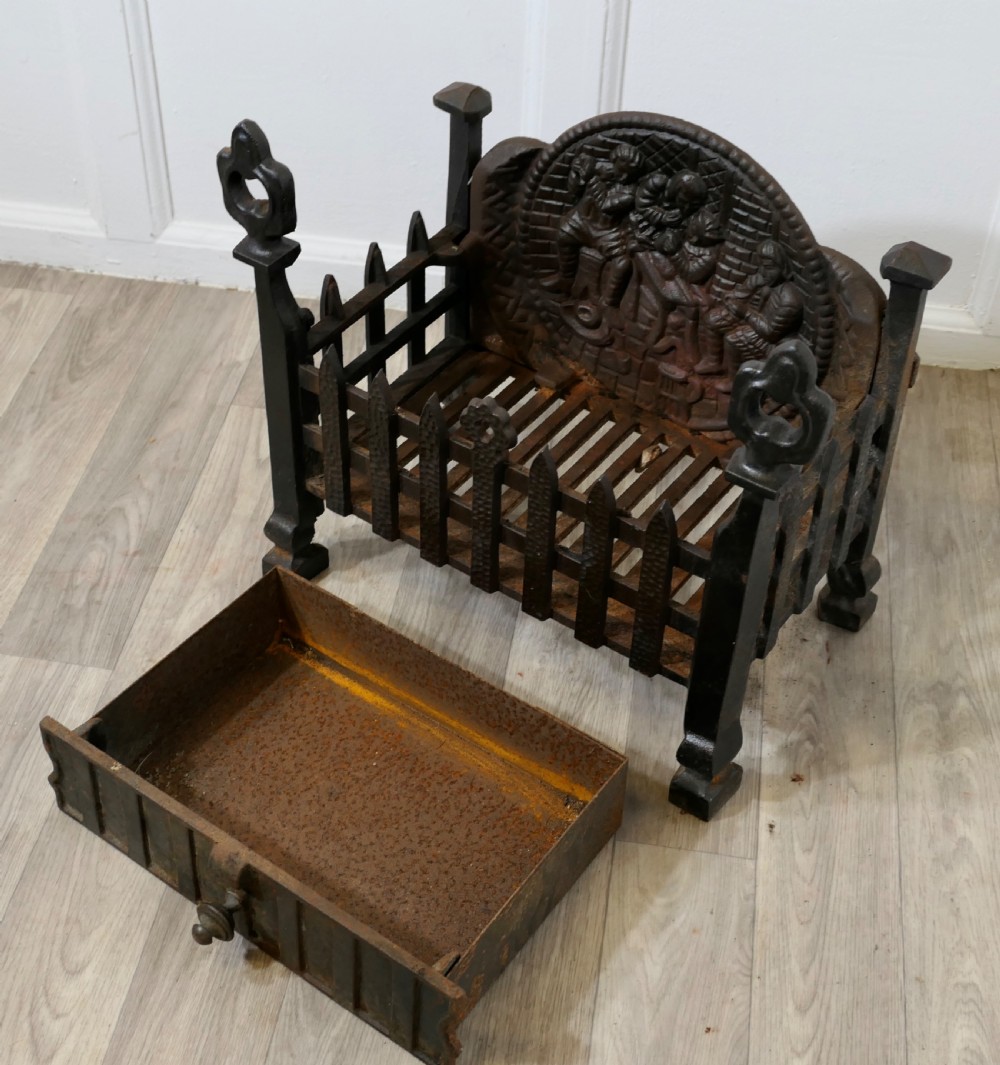 A heavy dark cast iron fire basket featuring a decorative relief backplate depicting figures, upright gothic finials, and a removable ash pan beneath. View V