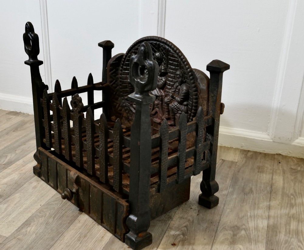 A heavy dark cast iron fire basket featuring a decorative relief backplate depicting figures, upright gothic finials, and a removable ash pan beneath. View VI