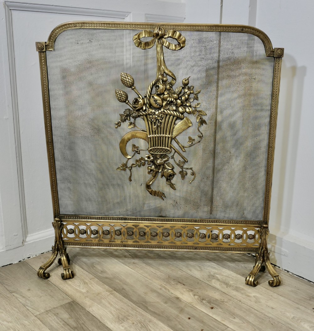 A freestanding brass fire guard featuring a central ribbon bow and floral basket relief mounted on fine wire mesh with scroll feet.