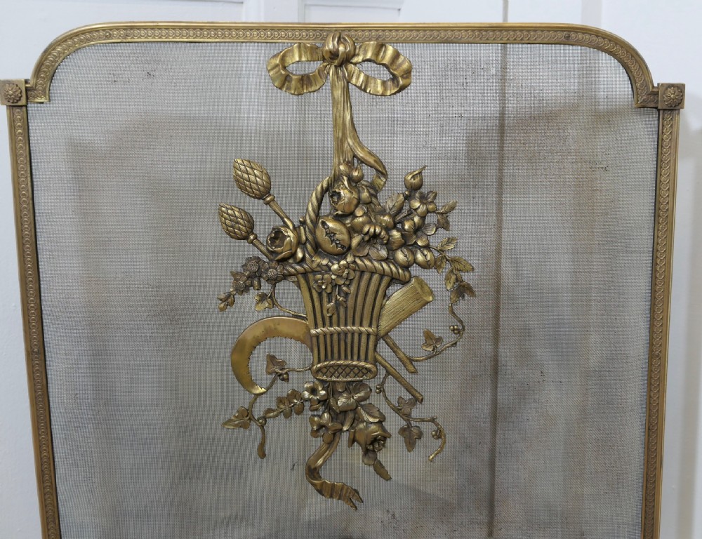 A freestanding brass fire guard featuring a central ribbon bow and floral basket relief mounted on fine wire mesh with scroll feet. View II