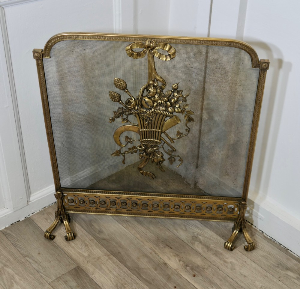 A freestanding brass fire guard featuring a central ribbon bow and floral basket relief mounted on fine wire mesh with scroll feet. View III