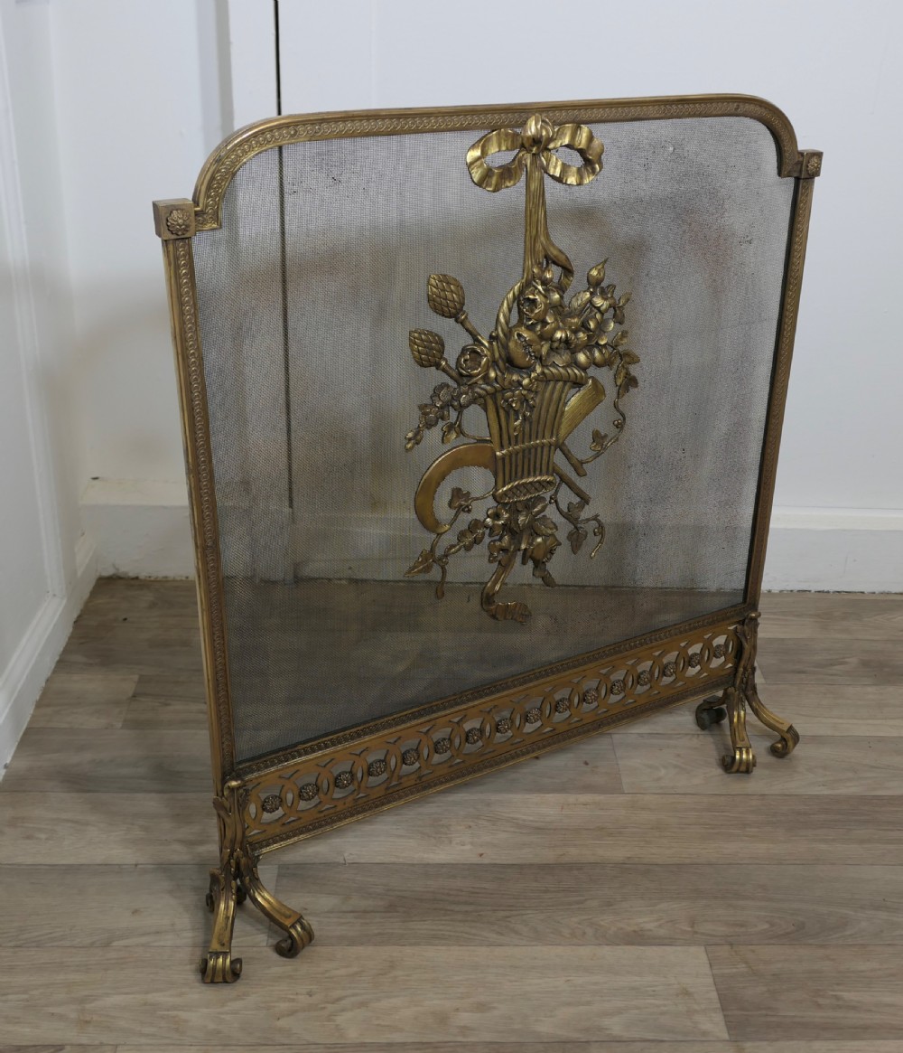 A freestanding brass fire guard featuring a central ribbon bow and floral basket relief mounted on fine wire mesh with scroll feet. View V
