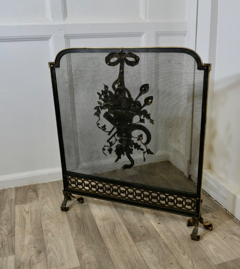 A freestanding brass fire guard featuring a central ribbon bow and floral basket relief mounted on fine wire mesh with scroll feet. View VI